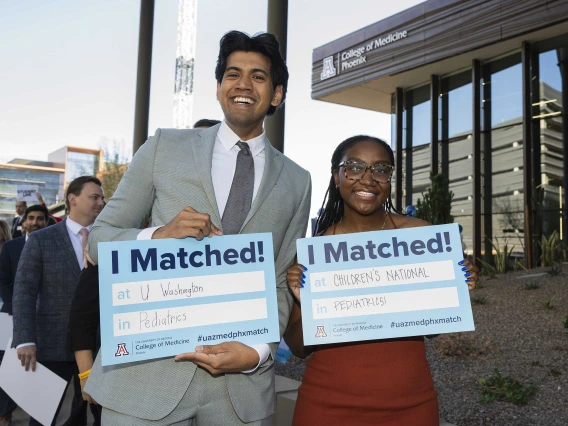 A young man and woman stand next to each other smiling and holding signs that read, “I matched!” in an outdoor setting. 