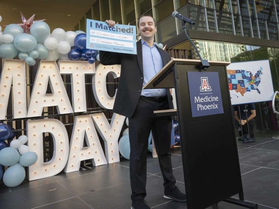 A medical student in a suit stands in front of a lectern holding up a small sign and smiling. 