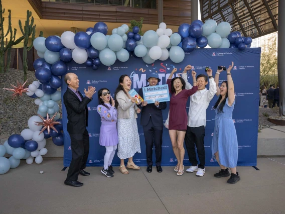 A group of seven cheering people stand in front of a blue background with balloons. 