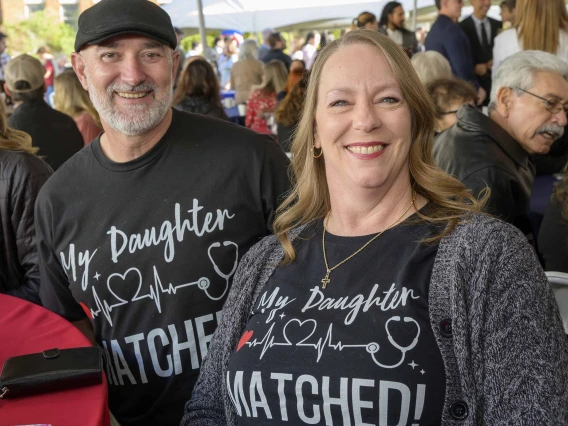 A man and woman sit at a table smiling with shirts that read, “My daughter matched!”