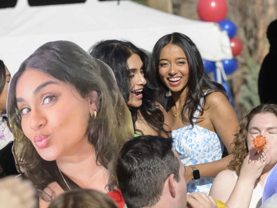 A large photo cutout of a woman’s face is held up in front of the same woman as she smiles and talks with another woman. 