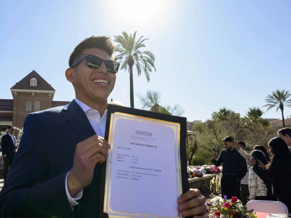 A young man in a suit and sunglasses smiles as he holds up a letter in an outdoor setting. 