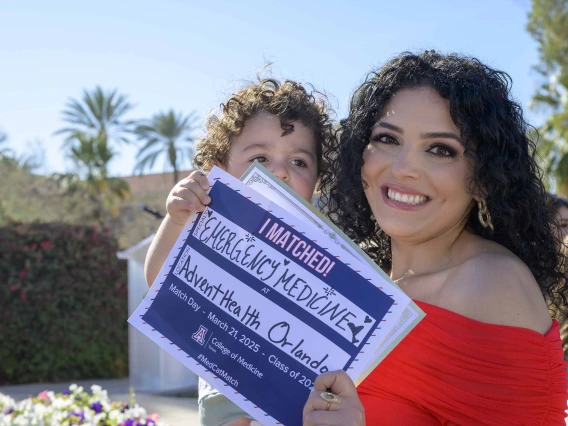 A woman in a red dress smiles as she holds a young boy and a sign that reads, “I matched!”.