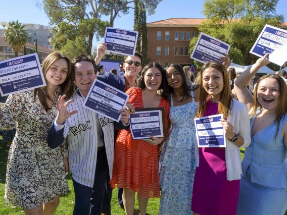 Seven well-dressed medical students stand outside holding small signs that announce where they will be going for their residencies. All are happy and smiling. 