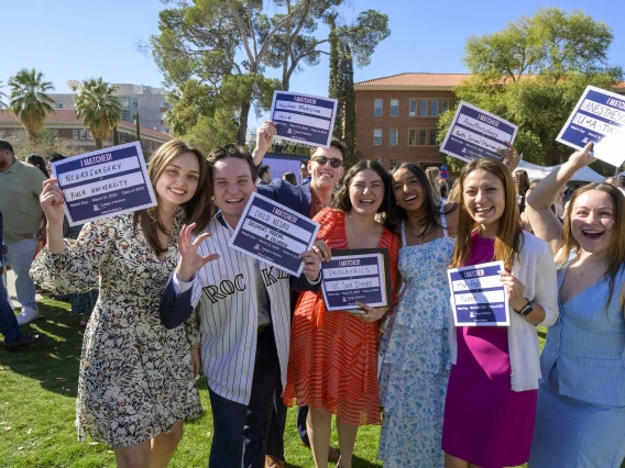 Seven well-dressed medical students stand outside holding small signs that announce where they will be going for their residencies. All are happy and smiling. 