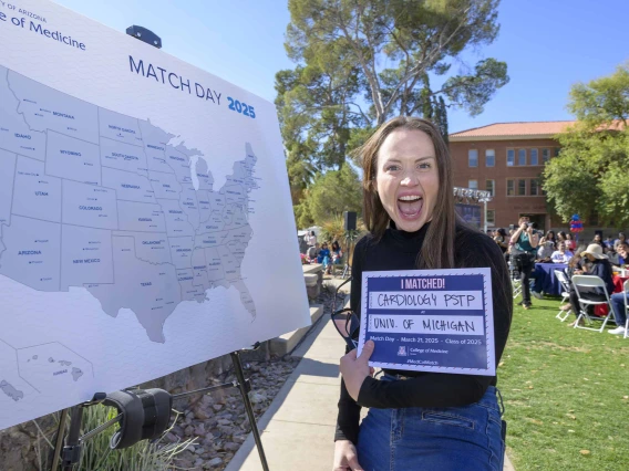 A woman holds up a piece of paper with writing on it noting that she will conduct her medical residency at the University of Michigan Hospital. She has an excited look on her face, with her mouth wide open. 