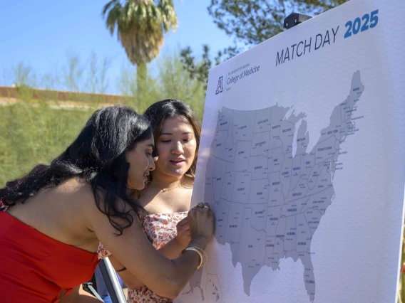 A young woman in a red dress puts a pin in a map of the United States while another young woman looks on. 