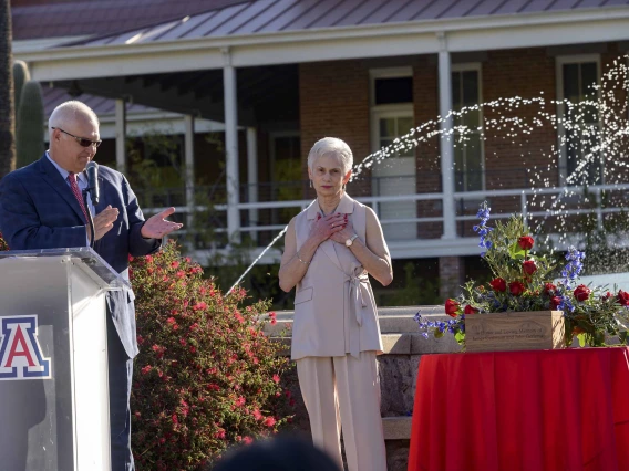 A man in a suit stands behind a lectern clapping as a woman stands nearby with her hands clasped to her heart. They are in an outdoor setting next to a table with flowers and a fountain in the background. 