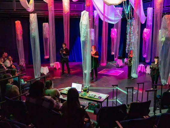 Five singers stand several feet apart on a stage with columns of translucent pink and blue mesh hanging from the ceiling between them