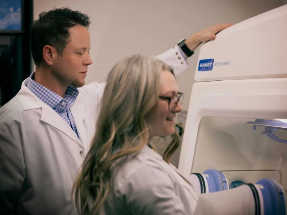 A researcher oversees a student using a glove box in a laboratory at the University of Arizona Cancer Center.