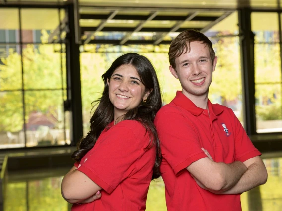 Two students standing back to back with their arms crossed in the lobby of the Health Sciences Innovation Building. 
