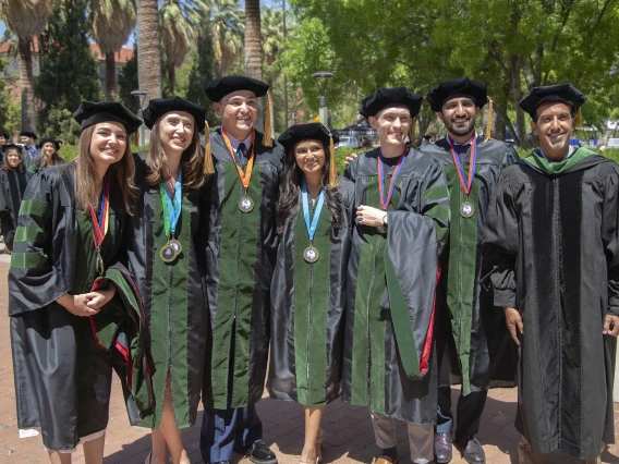 A group of graduating medical students poses for a photo on a sunny day outdoors.