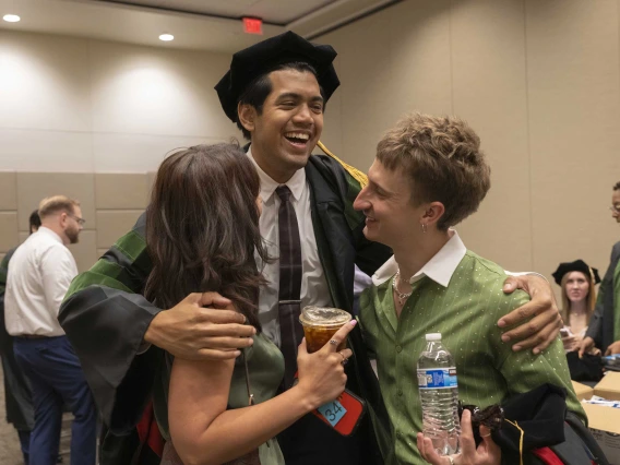 A tall young man wearing a graduation cap and gown smiles as he hugs two friends. 