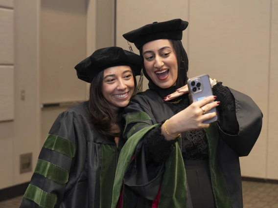 Two medical students in graduation caps and gowns smile as they take a selfie. 