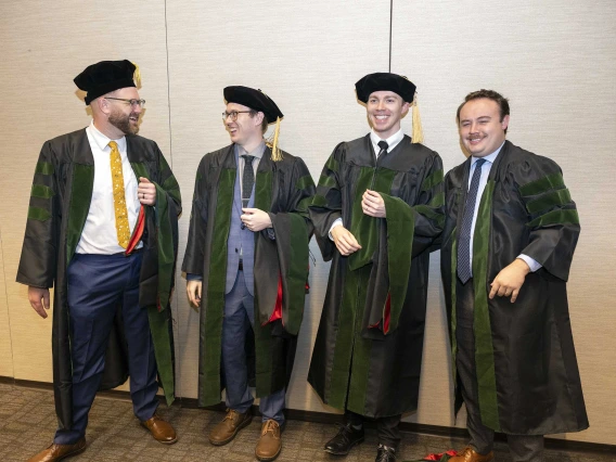 Four young men in graduation caps and gowns stand around, laughing. One of them has turned up his cap as a joke. 