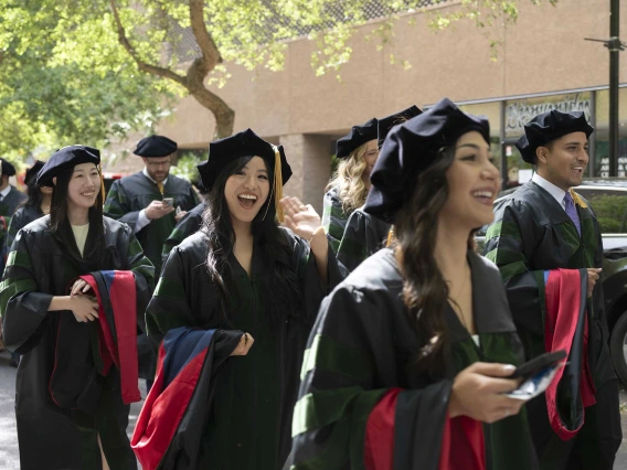 Several smiling young men and women in graduation caps and gowns walk in a line outside under some trees. 