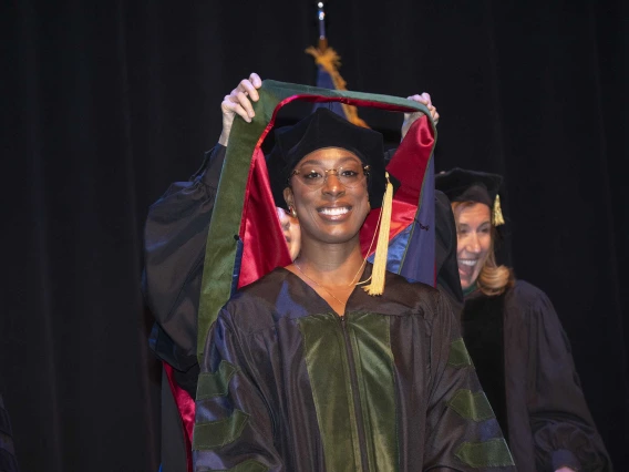A smiling woman wearing graduation regalia looks ahead as someone behind her places a ceremonial hood over her shoulders. 