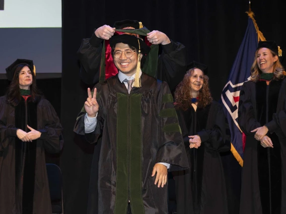 A young man wearing a graduation cap and gown smiles as he holds up two fingers in a “V” sign while a professor behind him places a ceremonial hood over his shoulders. 