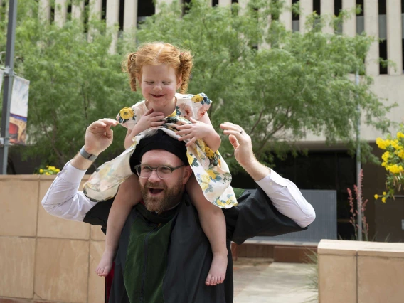 A bearded man wearing a graduation cap and gown smiles as his smiling young daughter sits on his shoulders. 
