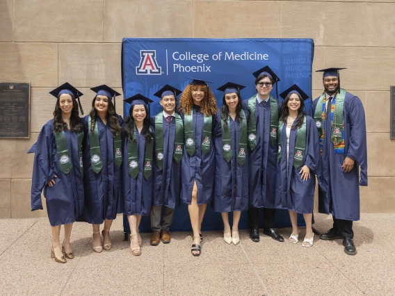 Nine students, all wearing graduation caps and gowns, stand together in front of a “College of Medicine – Phoenix” backdrop outside. 