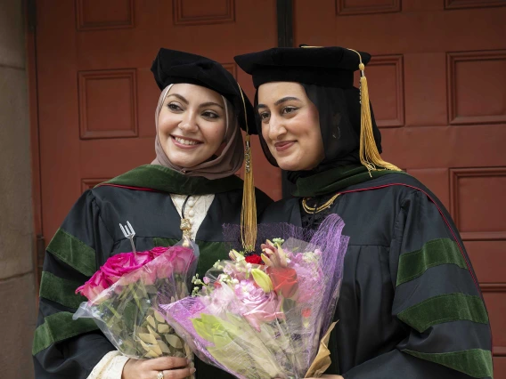 Two young women in graduation caps and gowns hold flowers and smile. 