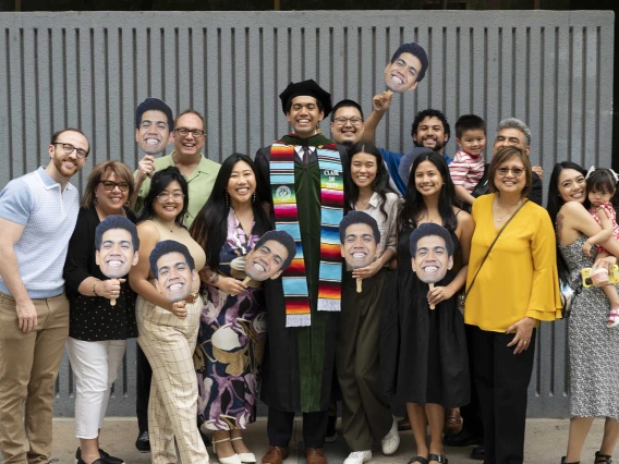 A tall, smiling young man in a graduation cap and gown stands with about a dozen family members, many of whom are holding photos of his face. 