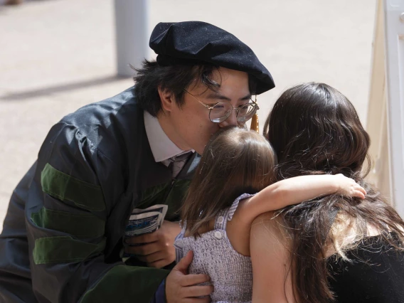 A man wearing a graduation cap and gown kisses his daughter, who is being held by his wife. 