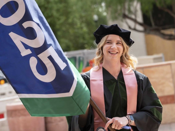 A young woman in graduation regalia smiles as she holds a large flag with “2025” on it. 