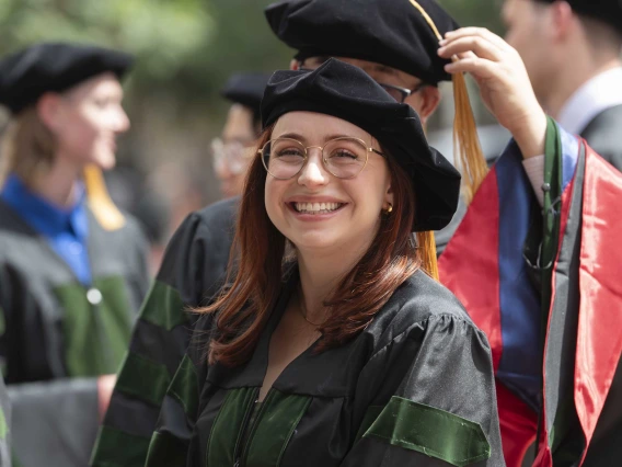 A young woman wearing graduation regalia smiles as other people dressed the same way are in the background. 