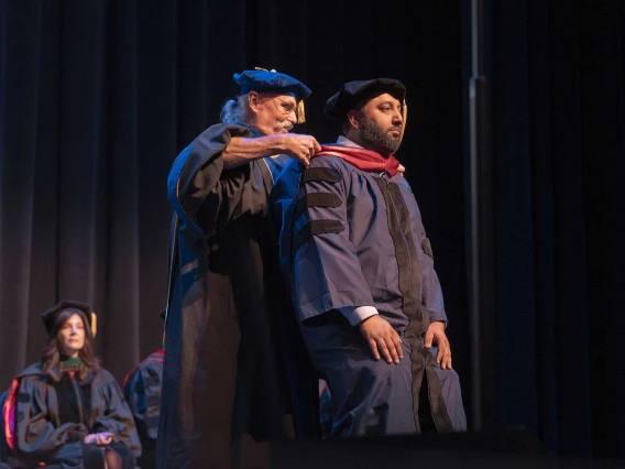 A professor places a ceremonial hood over the shoulders of a graduate onstage during a ceremony. Both are wearing graduation regalia. 