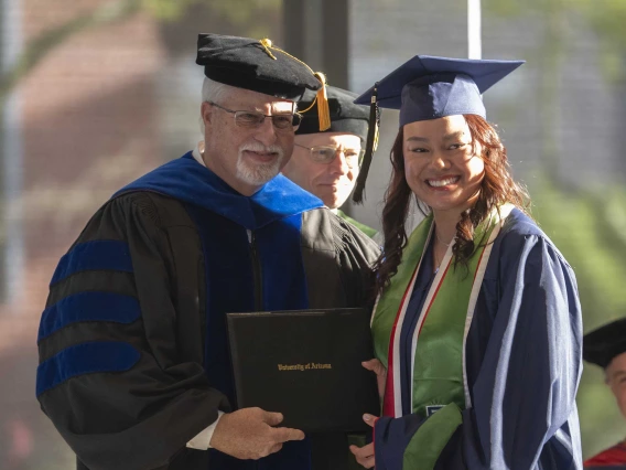 A male professor presents a diploma to a young woman. Both are smiling and wearing graduation regalia. 