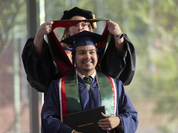 A young man wearing graduation regalia smiles as a professor standing behind him places a ceremonial hood on his shoulders.