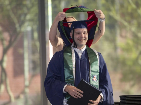 A young man in a graduation cap and gown smiles as a professor places a ceremonial hood over his shoulders. 