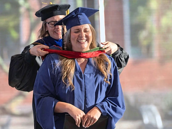 A professor places a ceremonial hood over the shoulders of a young woman. Both are smiling and wearing graduation regalia.