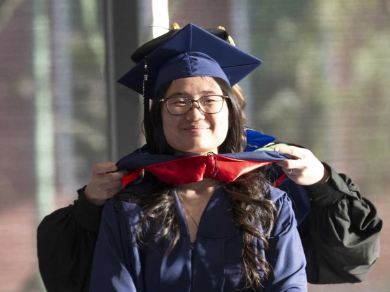 A young woman wearing graduation regalia smiles as a professor standing behind her places a ceremonial hood on her shoulders.