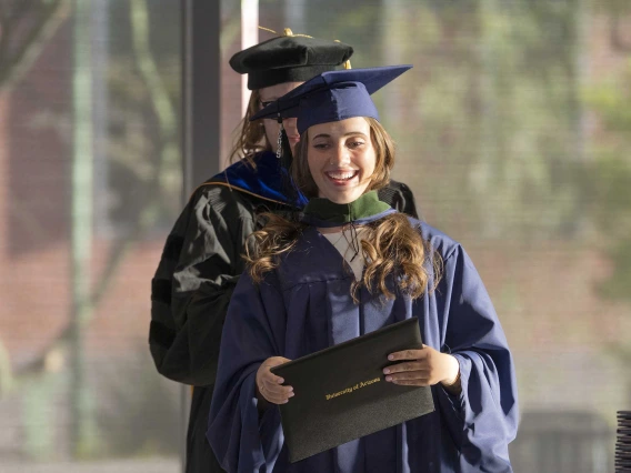 A young woman wearing graduation regalia smiles as she holds her diploma. An obscured professor stands behind her. 