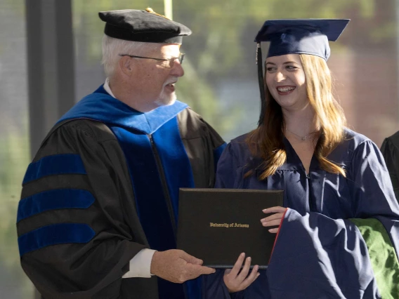 A male professor presents a diploma to a young woman. Both are smiling and wearing graduation regalia.