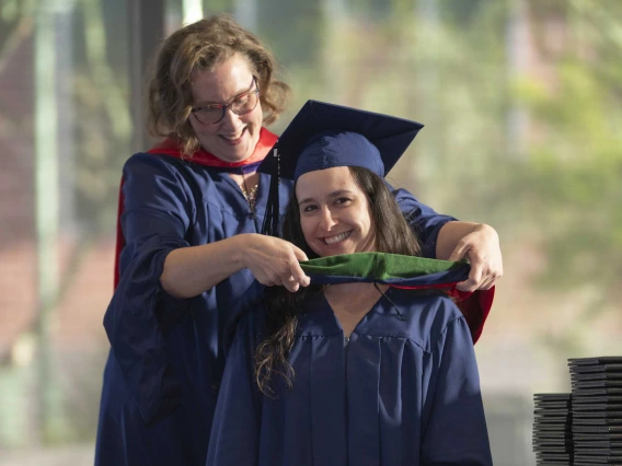 A professor places a ceremonial hood over the shoulders of a young woman. Both are smiling and wearing graduation regalia. 