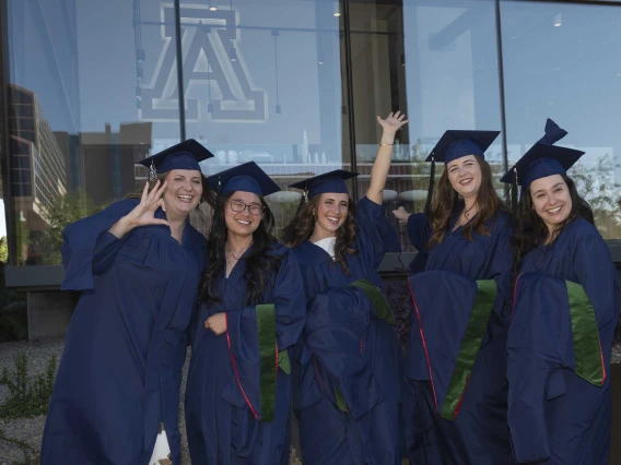 Five young women wearing graduation caps and gowns smile and cheer outside a glass building. 