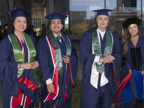 Four smiling graduates wearing caps and gowns stand outside a glass building. 