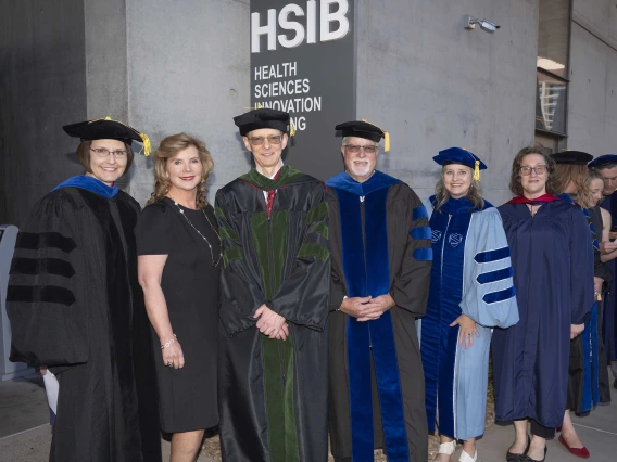 Six professors in graduation regalia stand together under a sign that reads “Health Sciences Innovation Building.”