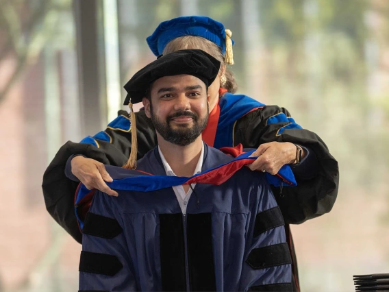 A student wearing a graduation cap and gown has a ceremonial hood placed over his shoulders by a professor. 