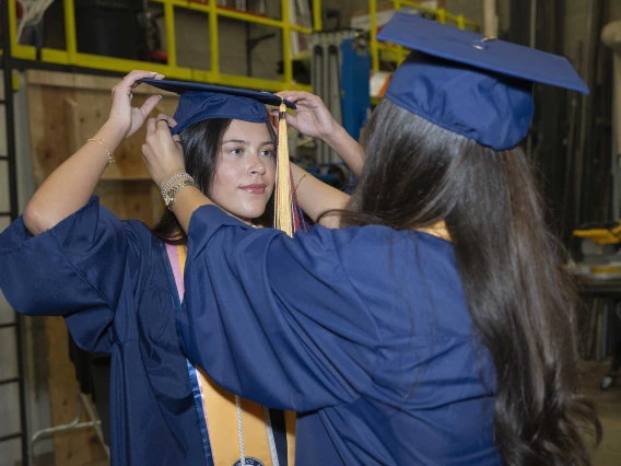 Two young women in graduation caps and gowns help each other set their mortarboards on straight. 