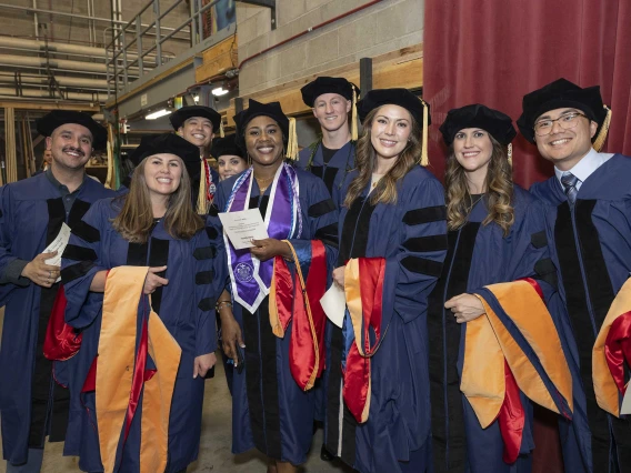 Nine College of Nursing students wearing caps and gowns stand backstage waiting for their graduation ceremony to begin. 