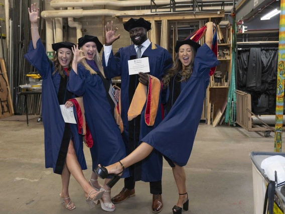 Four College of Nursing students wearing graduation caps and gowns wave and smile backstage before the graduation ceremony begins. 