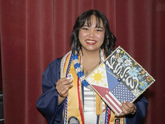 A smiling young woman wearing a graduation gown holds up her decorated mortar board and a “First Cat Grad” pin.