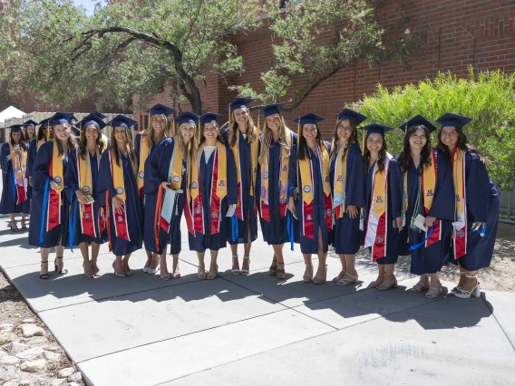 About a dozen nursing students, all dressed in graduation regalia, stand in a row outside a building. 