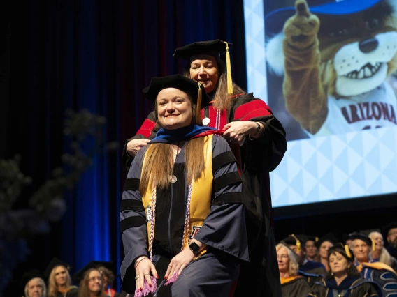 A smiling young woman wearing graduation regalia has a ceremonial graduation hood placed over her shoulders by a professor.