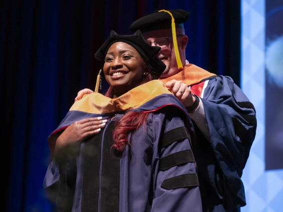 A smiling young woman wearing graduation regalia has a ceremonial graduation hood placed over her shoulders by a professor. 