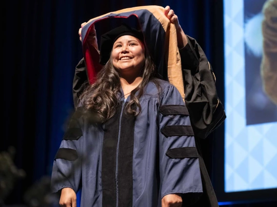 A smiling young woman wearing graduation regalia has a ceremonial graduation hood placed over her shoulders by a professor.